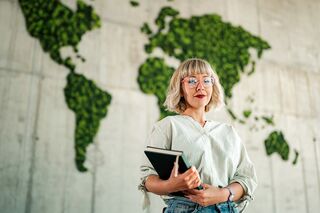 Junge Frau mit blonden Haaren und Brille steht mit Notebook in der Hand vor einer Wand mit einer moosbedeckten grünen Weltkarte