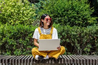 Eine Frau mit Sonnenbrille sitzt mit einem Laptop im Schneidersitz auf einer Bank.