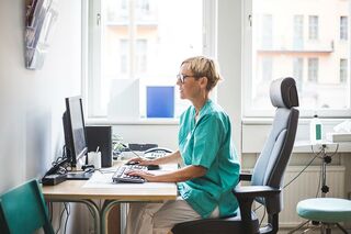Kurzhaarige Pflegebeschäftigte in Büro an Schreibtisch vor Computer.