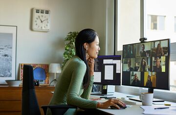 Junge Frau sitzt an Schreibtisch vor großem Monitor, auf dem andere Menschen zu sehen sind, und nimmt an einer Online-Lehrveranstaltung teil. 