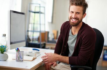 Junger Mann mit Bart sitzt an Schreibtisch und wendet sich lächelnd zur Kamera