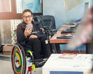 Junge Frau im Rollstuhl sitzt lächeln an Schreibtisch und blickt auf Smartphone
