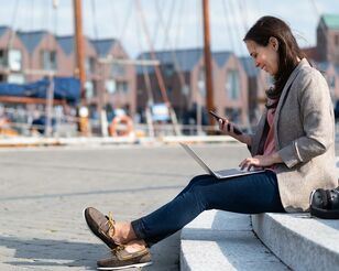 Junge Frau sitzt auf Treppenstufen am Hafen mit Notebook und Smartphone