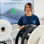 Züleyha Saraç, wissenschaftliche Mitarbeiterin, steht lächelnd vor einem Screen mit technischen Zeichnungen, im Vordergrund sind technische Geräte einer Werkstatt zu sehen.