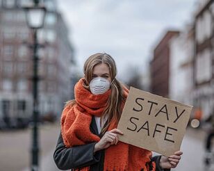 Blonde Frau mit rotem Schal steht draußen mit einem Pappschild auf dem Stay safe steht. 