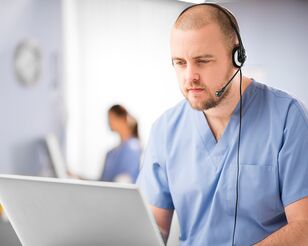 Männliche Pflegekraft sitzt mit Headset vor Notebook.