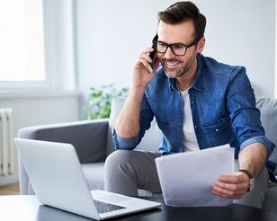 Ein Mann sitzt auf dem Sofa, telefoniert, schaut auf einen Laptop und hält Papiere in der Hand.