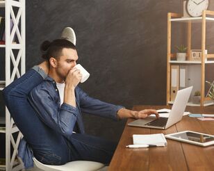 Mann sitzt vor einem Laptop, trinkt Kaffee und macht Yoga