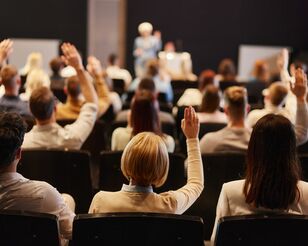 Viele Menschen in Rückansicht sitzen auf einer Internationalen Konferenz und hören einen Vortrag. Einige heben die Hände, um Fragen zu stellen.