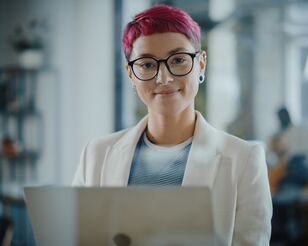 Eine Person hält einen geöffneten Laptop und schaut lächelnd in die Kamera.