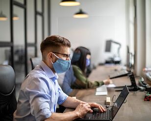 Zwei Personen mit Corona-Masken sitzen an Schreibtisch und arbeiten mit Notebooks