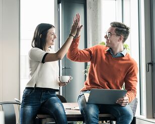 Junge Frau und Mann sitzen auf Tisch, freuen sich und geben High Five.