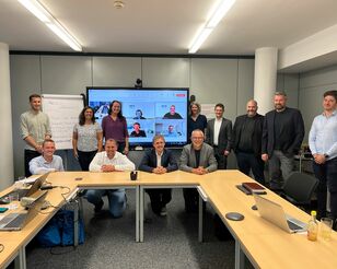 Menschen sitzen und stehen um einen Tisch herum und lächeln in die Kamera. Im Hintergrund ist ein großer Bildschirm, auf dem weitere Menschen iun einer digitalen Konferenz zugeschaltet sind.