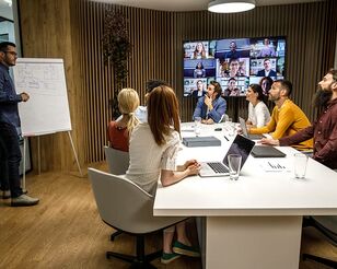 Lernsituation in einem Lehrraum mit Personen an Tisch und zugeschalteten Online-Teilnehmenden auf großem Screen.