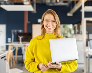 Eine Frau mit Laptop im Arm lächelt in die Kamera.