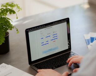 Laptop auf Schreibtisch mit Unterlagen daneben und halbsichtbarer sitzender Person