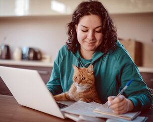 Dunkelhaarige Frau an Schreibtisch mit Notebook macht Notizen auf Papier und hat eine Katze auf dem Schoß.