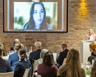 Blick in die Veranstaltung zur Verleihung des Studienpreises 2025 mit sitzenden Gästen, einer Rednerin an Sprechpult und der Preisträgerin auf einem Screen an der Wand.