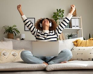 Eine Person mit Kopfhörern und Laptop auf dem Schoß sitzt auf Sofa und reckt beide Arme jubelnd nach oben.
