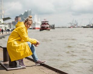 Blonde Frau im gelben Regenmantel sitzt an den Landungsbrücken am Wasser und blickt in die Kamera; im Hintergrund Schiffe und die Elbphilharmonie.