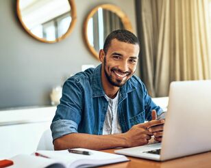 Ein Mann sitzt in seinem Wohnzimmer am Tisch vor einem Laptop und lächelt in die Kamera.