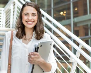 Junge Frau mit Unterlagen und Notebook in der Hand steht auf einer Treppe und lächelt in die Kamera.