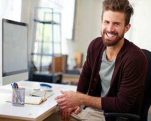 Junger Mann mit Bart sitzt an Schreibtisch und wendet sich lächelnd zur Kamera