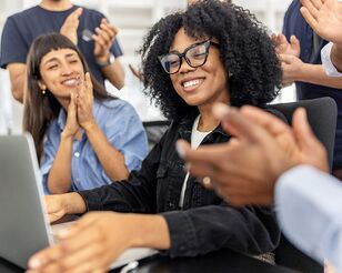 junge Frau mit Brille sitzt mit zufriedenem Blick vor Laptop und erhält Applaus von umstehenden Personen