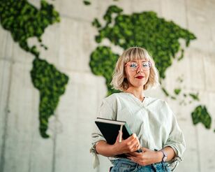 Junge Frau mit blonden Haaren und Brille steht mit Notebook in der Hand vor einer Wand mit einer moosbedeckten grünen Weltkarte