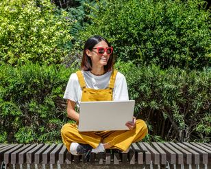 Eine Frau mit Sonnenbrille sitzt mit einem Laptop im Schneidersitz auf einer Bank.