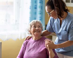 junge Pflegerin hält Seniorin im Rollstuhl die Hand, beide blicken sich lächelnd an