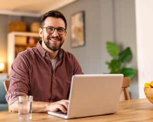 Ein Mann sitzt vor einem Laptop an einem Tisch und lächelt in die Kamera.