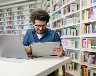 Lockiger Mann sitzt in an Schreibtisch vor Bücherregalen und hantiert mit Laptop und Tablet.