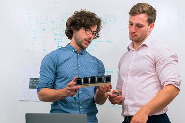 Zwei Männer betrachten Bauteil vor Whiteboard