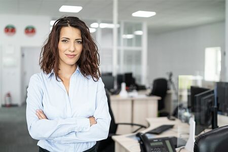 Frau mit verschränkten Armen in hellblauer Bluse steht in einem modernen Büro mit Schreibtischen und Computern im Hintergrund.