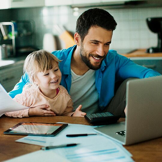 Vater und Tochter am Küchentisch. Vater hat Laptop aufgeklappt und Lernmaterialien auf dem Tisch.