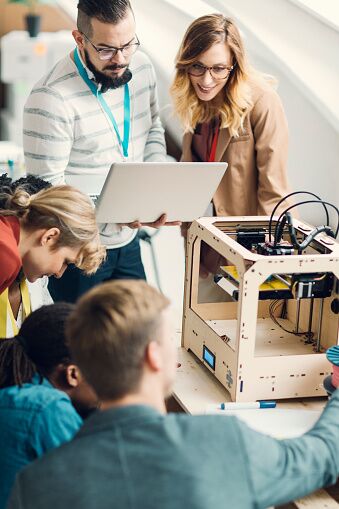 Das Bild zeigt eine Gruppe von Studenten, die um einen Tisch sitzen, auf dem ein 3D-Drucker steht. 