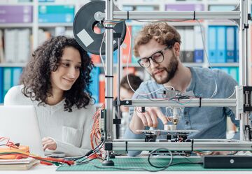HFH Bachelorstudiengang Digital Engineering Ein Mann und eine Frau gucken sitzen vor einem 3D Drucker und gucken ihm beim Drucken eines Bauteils zu
