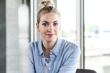 HFH Bachelor Wirtschaftspsychologie Blonde Frau mit blauem Hemd sitzt im Büro und lächelt in die Kamera