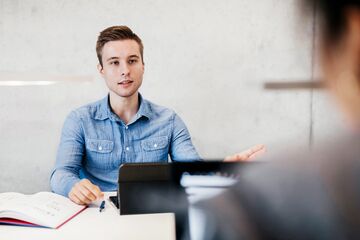 HFH Master Management im Gesundheitswesen Mann mit blauem Hemd sitzt im Büro und unterhält sich mit anderer Person
