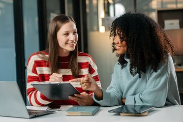 Zwei junge Frauen sitzen an einem Schreibtisch mit Notebook und betrachten gemeinsam ein Tablet.