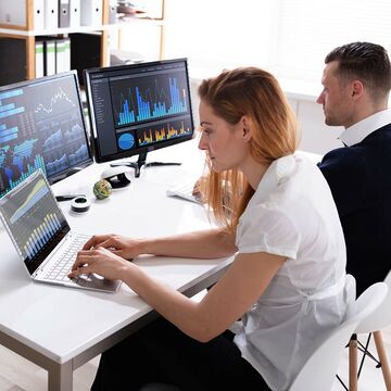 Frau und Mann sitzen nebeneinander an Schreibtisch mit zwei Bildschirmen sowie einem Notebook und arbeiten.
