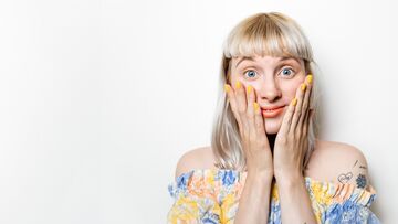 Blonde, junge Frau wirft überrascht die Hände vors Gesicht.