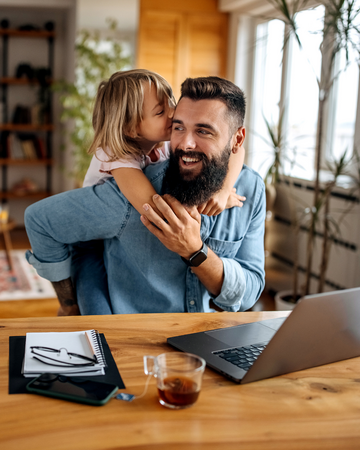 Vater sitzt am häuslichen Schreibtisch vor einem Laptop und wird von hinten von seiner kleinen Tochter umarmt