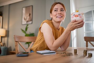Eine rothaarige Frau sitz lächelnd an einem Tisch, in der Hand ein Sparschwein.