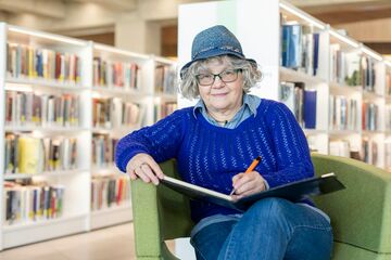 Eine älztere FRrau sitzt in einer Bibliothek in einem Sessel und schreibt in ein Heft, sie lächelt in die Kamera.